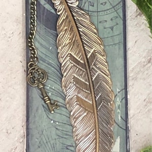 Puede incluir: Un marcapáginas de metal dorado con forma de pluma con un dije de llave unido a una cadena. El marcapáginas está sobre un fondo con un diseño verde y negro.