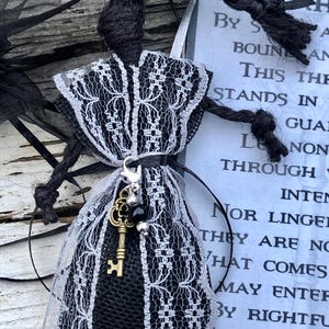 Może przedstawiać: Ręcznie robiona czarno-biała lalka z warkoczem, ubrana w sukienkę z białym koronkowym wykończeniem. Mały brelok w kształcie klucza wisi u talii lalki. W tle widoczna jest etykieta z tekstem.