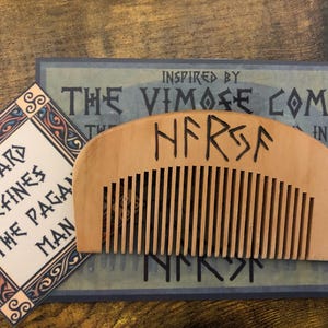 Op de afbeelding: Een ronde houten baardkam met een rij tanden. De kam is gegraveerd met het woord "NFRSA" in runenschrift. De kam ligt op een oppervlak met een achtergrondafbeelding van een baardkam met de tekst "Inspired by The Vimose Comb".