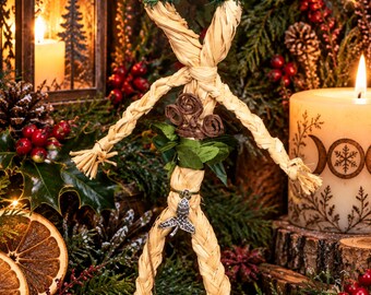 May include: A straw figure crafted from braided natural fibers, featuring a dark brown coiled headpiece, red berry accents, green foliage at the waist, and a small silver-toned metal charm shaped like a leaf hanging from the center.