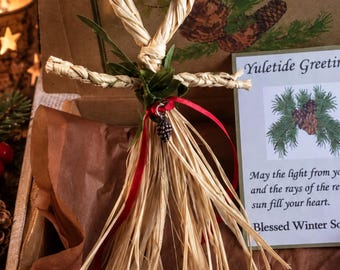 Könnte beinhalten: Eine Strohpuppe als Ornament mit einem geflochtenen Schlaufenkopf, ausgestreckten Armen und einem Rock aus losen Strohhalmen. Sie ist mit kleinen braunen Holzrosen, grünen Blättern, roten Beeren, einem roten Band und einem Metallanhänger verziert.