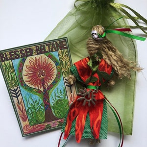 May include: A handmade doll with a green and orange dress, a brown hair, and a silver sun charm. The doll is sitting on a green fabric bag with the text "Blessed Beltane" on a card.