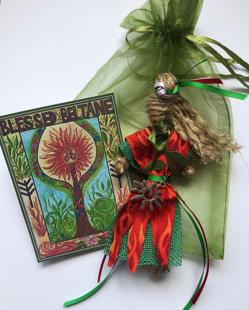May include: A handmade doll with a green and orange dress, a brown hair, and a silver sun charm. The doll is sitting on a green fabric bag with the text "Blessed Beltane" on a card.