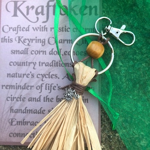 May include: A small, brown, woven corn husk doll keychain with a wooden bead and a silver charm. The keychain is attached to a silver key ring. The doll is hanging from a card with the text "Kraflaken Crafted with rustic charm this Keyring Charm small corn doll, echoes country traditions nature's cycles. A reminder of life's circle and the beauty in handmade simplicity Embrace connection".