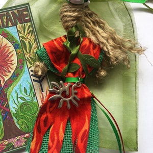May include: A handmade doll with a long, brown, string hair, wearing a red and green dress with a silver sun pendant. The doll is sitting in a green mesh bag with red and green ribbons.