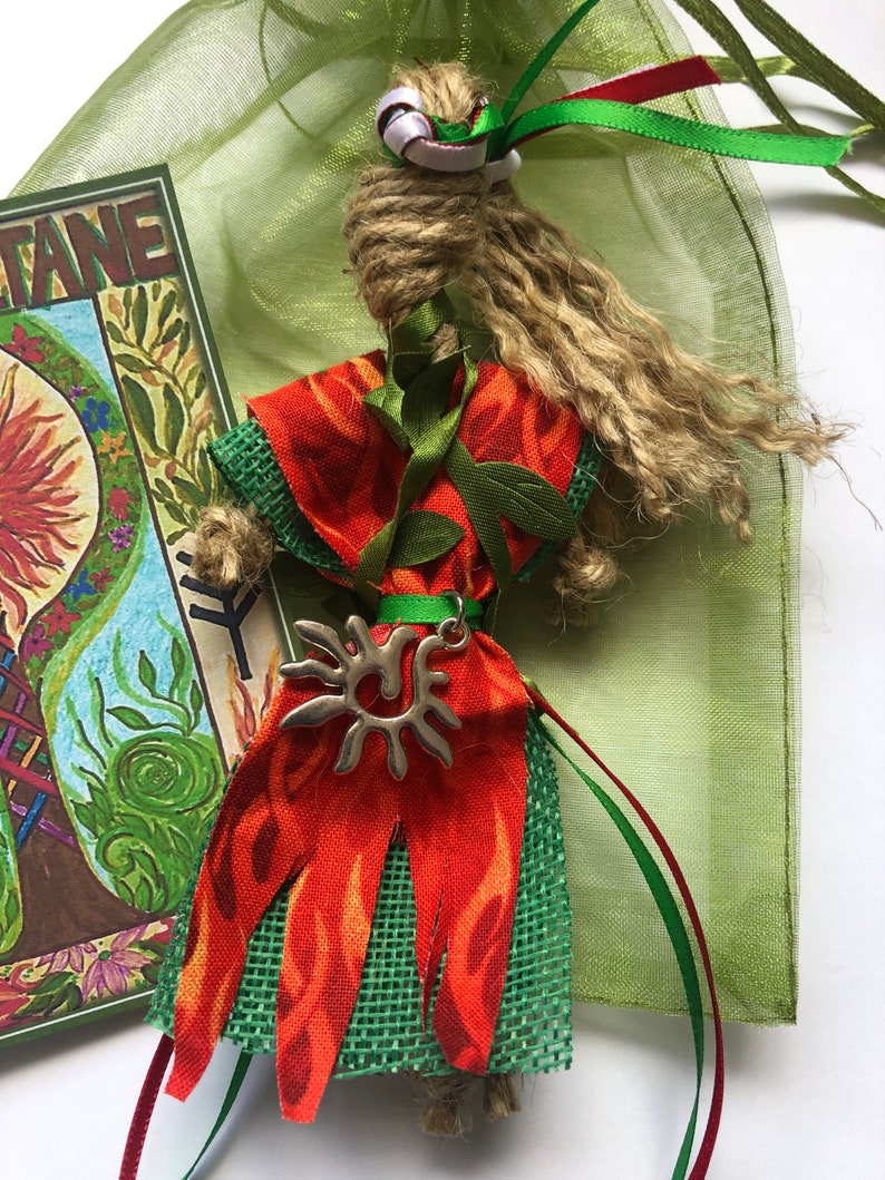 May include: A handmade doll with a long, brown, string hair, wearing a red and green dress with a silver sun pendant. The doll is sitting in a green mesh bag with red and green ribbons.