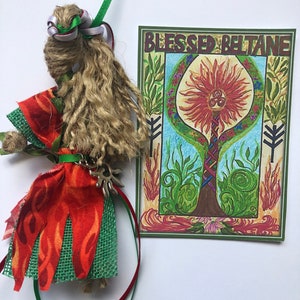 May include: A handmade doll with long brown hair, wearing a red and green dress with a silver charm. The doll is next to a card with the words "Blessed Beltane" and a drawing of a tree with a sun and a triple spiral symbol.