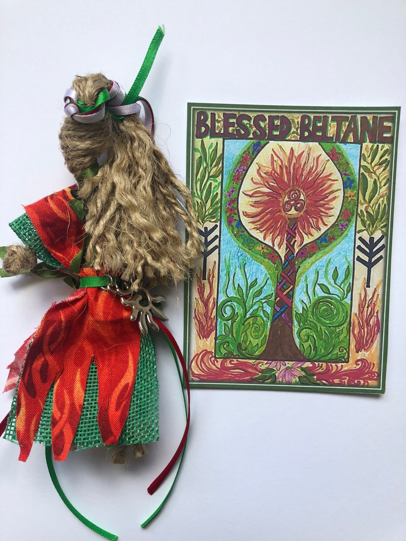 May include: A handmade doll with long brown hair, wearing a red and green dress with a silver charm. The doll is next to a card with the words "Blessed Beltane" and a drawing of a tree with a sun and a triple spiral symbol.