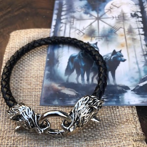 Può includere: Braccialetto in pelle nera con due chiusure a testa di lupo. Il braccialetto è intrecciato e ha una finitura argentata. Il braccialetto è su uno sfondo di juta.