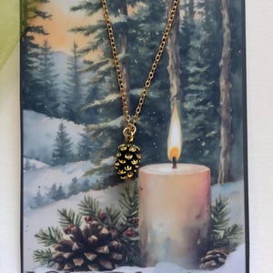 May include: A gold-colored chain necklace with a pinecone pendant hangs in front of a winter-themed greeting card. The card features a snowy forest scene, a lit candle, and the words "Yule Blessings".