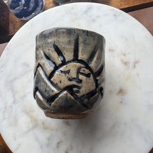 May include: Handmade ceramic cup with a sun and mountain design. The cup is primarily tan with dark blue accents, featuring a smiling sun and stylized mountain range. The cup is sitting on a white marble surface.