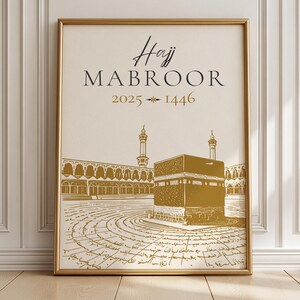 Peut inclure: Une affiche encadrée sur fond crème avec le texte noir Hajj MABROOR et le texte doré 2025 1446. Sous le texte se trouve un dessin au trait doré de la Kaaba et de l'architecture de la mosquée environnante sur un sol carrelé.
