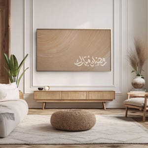 Puede incluir: Una obra de arte enmarcada con un diseño abstracto beige y caligrafía árabe blanca. La obra cuelga sobre un mueble de madera en un salón. La habitación presenta una paleta de colores neutros, un otomán tejido y una alfombra estampada.