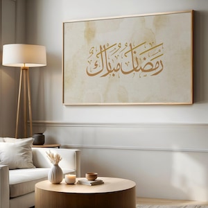 May include: A rectangular wall art piece featuring gold Arabic calligraphy on a textured, cream-colored background with subtle tan watercolor-style splotches. The artwork is enclosed in a thin, metallic gold frame and displayed on a wall.