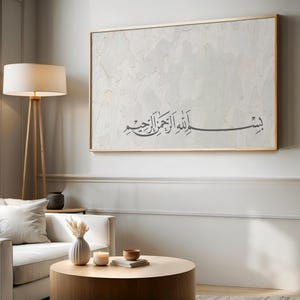 May include: A rectangular wall art piece featuring a light, textured, off-white background with a thin gold frame. The artwork displays dark gray Arabic calligraphy in the center, which reads "Bismillah ar-Rahman ar-Rahim" in a flowing script.