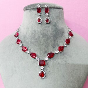 May include: A red and silver jewellery set, comprising a necklace and matching earrings, displayed on a grey mannequin. The necklace features red teardrop-shaped stones and a large square stone at the centre. The earrings have a rectangular top and a teardrop-shaped bottom.