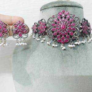 May include: A silver-toned choker necklace and matching earrings set. The jewellery features a floral design with vibrant red gemstones and small white bead accents. The necklace is displayed on a grey mannequin head.