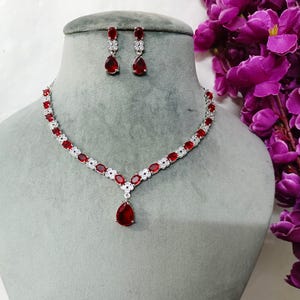 Peut inclure: Un collier en argent avec un pendentif en forme de larme rouge et des boucles d'oreilles assorties. Le collier est orné de petites pierres claires.