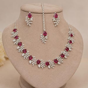 May include: A jewelry set consisting of a necklace, a pair of earrings, and a maang tikka. Each piece features silver-toned metal settings adorned with clear white stones and teardrop-shaped deep pink stones arranged in a leaf-like pattern.