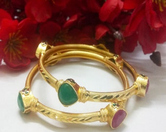 May include: Two gold bangles with green and red gemstones. The bangles are decorated with a chevron pattern.