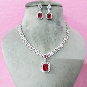 Puede incluir: Un conjunto de joyas plateadas con un collar y pendientes a juego. El collar tiene un colgante con una gran gema roja rectangular rodeada de piedras blancas más pequeñas. Los pendientes también tienen gemas rojas rectangulares.