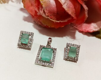 Könnte beinhalten: Ein Set aus silbernem Schmuck mit einem Anhänger aus grünem Edelstein und passenden Ohrringen. Anhänger und Ohrringe haben einen quadratischen Edelstein, der von einem silbernen Rahmen mit kleinen, klaren Steinen umgeben ist.
