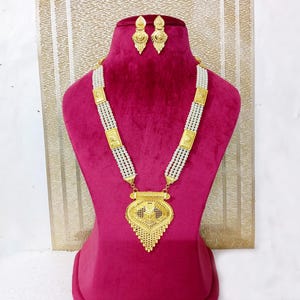 May include: A gold necklace with a large, ornate pendant and a matching pair of earrings. The necklace is made of multiple strands of white beads and features gold accents.