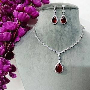 以下が含まれることがあります： 赤い宝石がセットされた涙型のペンダントが付いたシルバーネックレス。ネックレスには、赤い宝石の涙型のダングルが付いたマッチングイヤリングが付属しています。