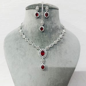 May include: A silver-coloured jewellery set comprising a necklace, earrings, and a headpiece. The set features oval, deep red gemstones set in silver metal. The necklace and headpiece have a floral design. The jewellery is displayed on a grey mannequin head.