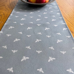 May include: A grey table runner with a white bee pattern. The runner is on a wooden table with a bowl of apples on top.