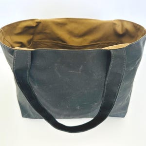 May include: A dark charcoal grey tote bag with two matching shoulder straps. The interior of the bag is lined with a contrasting tan fabric and features an open pocket along the back wall. The exterior has a matte, textured finish.