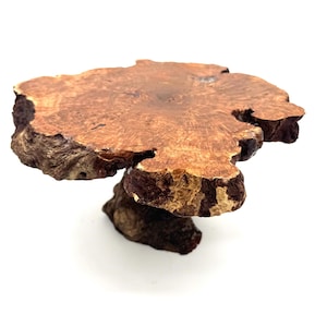 May include: A natural wood pedestal with a unique, organic shape. The tabletop is a rich brown color with a smooth surface, supported by a textured, dark brown base. The edges have a rough, bark-like appearance.