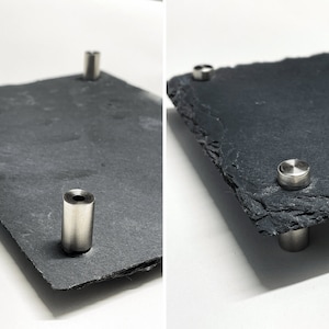 Puede incluir: Una bandeja de servir de pizarra negra con cuatro soportes de metal plateado. La bandeja tiene una textura rugosa y natural.