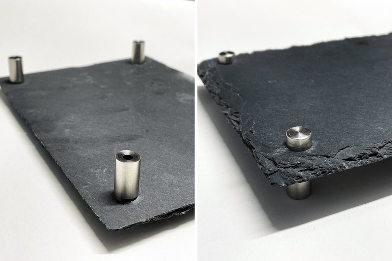 Puede incluir: Una bandeja de servir de pizarra negra con cuatro soportes de metal plateado. La bandeja tiene una textura rugosa y natural.