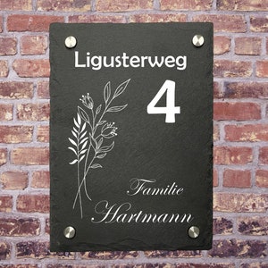 May include: Black slate house number sign with white lettering. The sign reads "Ligusterweg 4 Familie Hartmann".