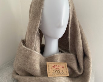 May include: A beige alpaca infinity scarf displayed on a mannequin head. The scarf is soft and fluffy, with a looped design. A small tag reads "Alpaca Yummy's Boutique". The scarf is a neutral color, suitable for various outfits.