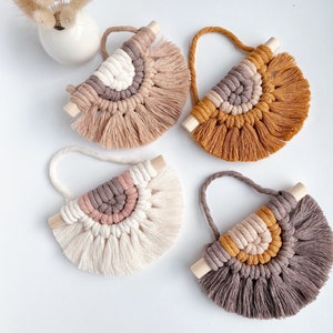 Könnte beinhalten: Vier Makramee-Wandbehänge in neutralen Farben, darunter Beige, Creme und Braun, mit Holzstäben und Aufhängeschlaufen. Die fächerförmigen Designs haben einen strukturierten, gefransten Boden. Eine kleine weiße Vase mit Trockenblumen befindet sich im Hintergrund.