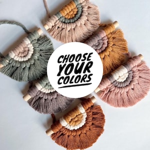 May include: A collection of handmade macrame wall hangings in various colors, including muted greens, browns, and pinks. Each piece features a half-circle design with a wooden dowel and a looped handle. The text "CHOOSE YOUR COLORS" is in the center.