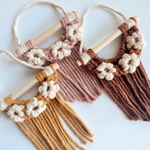 Könnte beinhalten: Drei Makramee-Wandbehänge mit Gänseblümchen-Akzenten. Jedes hat einen Holzstab, eine Schlaufe zum Aufhängen und einen Fransen. Die Farben sind Creme, Rosa, Braun und Senfgelb, mit weißen Gänseblümchen-Details.