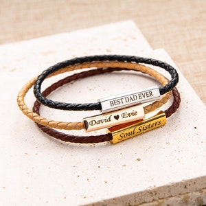May include: Three braided leather bracelets in black, tan, and brown, each with a metal bar. The bars are engraved with the phrases "BEST DAD EVER", "David ♥ Evie", and "Soul Sisters". The bracelets are displayed on a textured, off-white surface.