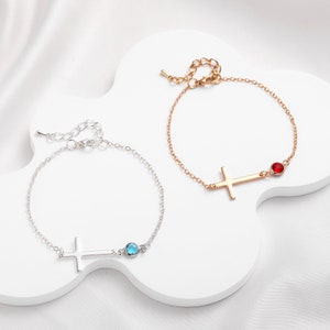 May include: Two delicate cross bracelets with small gemstone accents. One bracelet features a blue gemstone, while the other has a red gemstone.