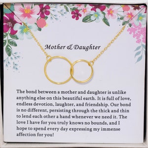 May include: A gold necklace with two interlocking circles, presented in a white box with a floral design. The box includes a message about the bond between a mother and daughter. "Mother & Daughter" is written at the top of the box.