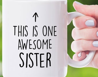 Puede incluir: Taza de cerámica blanca con texto negro que dice "This is one awesome sister" con una flecha apuntando hacia arriba.