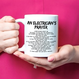 May include: White ceramic mug with the text "AN ELECTRICIAN'S PRAYER" in black. The prayer is printed in a black, easy-to-read font. The mug is held by a person with pink nail polish.