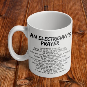 May include: White ceramic mug with the text "An Electrician's Prayer" in black. The prayer is printed in a black, stylized font. The mug has a curved handle and is set against a wooden background.