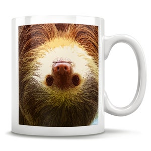 May include: A white ceramic coffee mug with a brown and tan sloth face printed on it. The sloth is upside down and looking directly at the viewer.