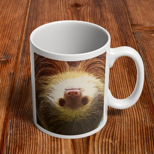 May include: White ceramic coffee mug with a brown and white sloth hanging upside down on the front. The sloth's face is visible, with a pink nose and black eyes.
