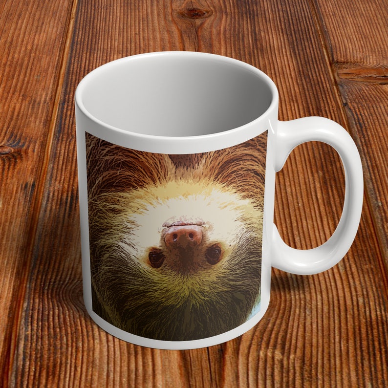 May include: White ceramic coffee mug with a brown and white sloth hanging upside down on the front. The sloth's face is visible, with a pink nose and black eyes.