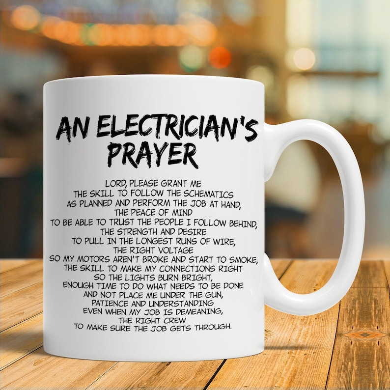May include: White ceramic mug with a large handle. The mug features the text "AN ELECTRICIAN'S PRAYER" in black, along with a prayer for electricians. The mug is on a wooden surface.