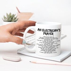 May include: White ceramic mug with the text "An Electrician's Prayer" in black. The mug features a list of aspirations for an electrician. The mug is held by a person with light blue nail polish.